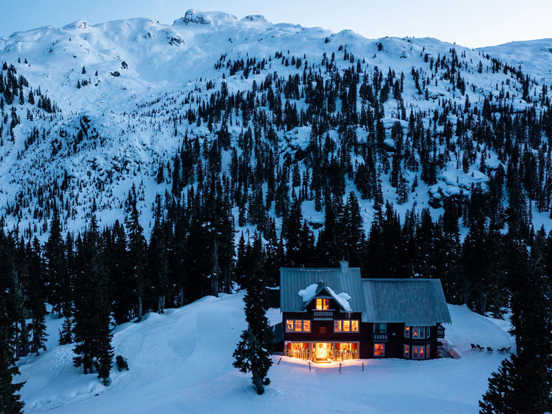 A warmly lit cabin surrounded by snowy trees and mountains